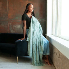 A woman sits on a couch draped in the Sorbet Mist Blanket, showcasing its soft green hue and cozy texture.