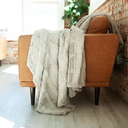 Beige gray blanket draped over an brown chair in a room with a brick wall and plant.