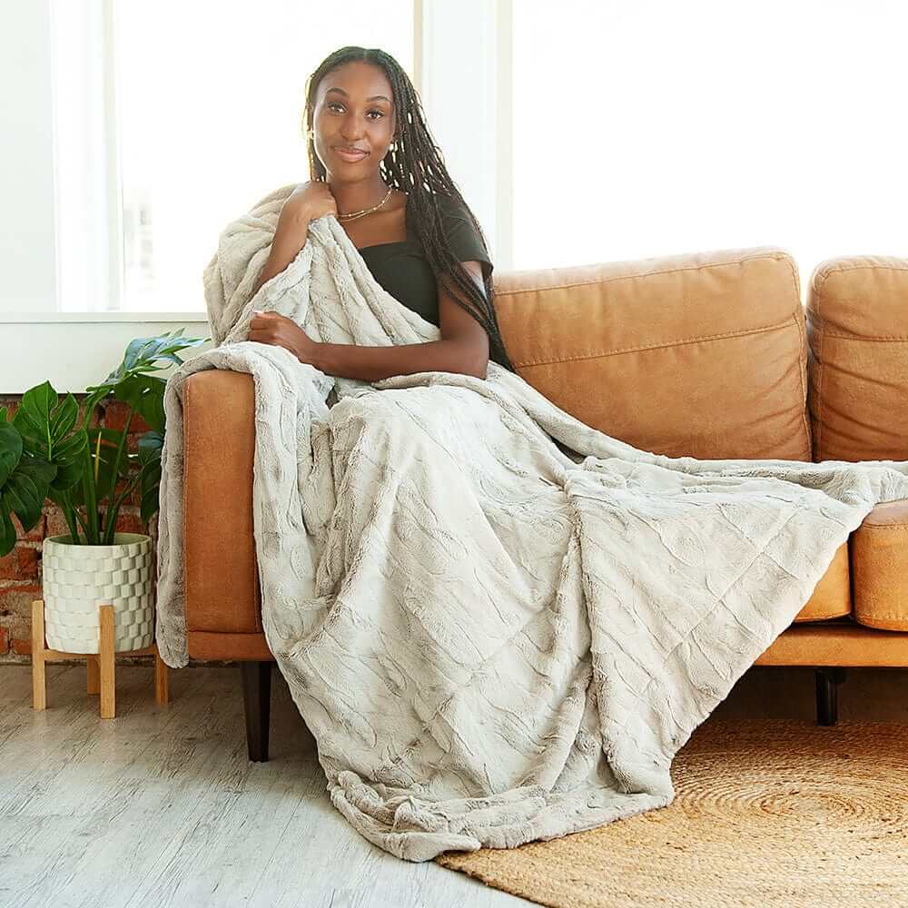 Woman sitting on a brown couch with a beige soft blanket draped over her, in a bright room with plants.