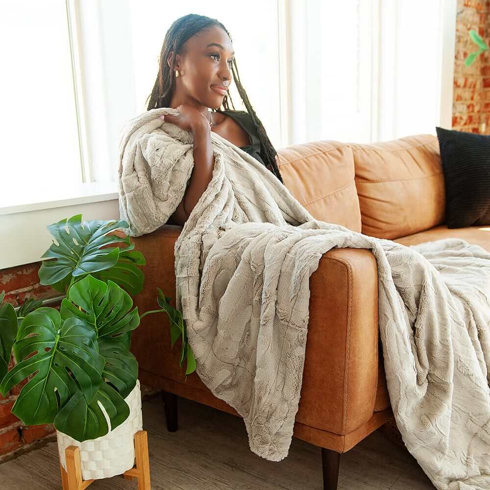 Woman sitting on a couch with a grayblanket, surrounded by plants in a bright room.