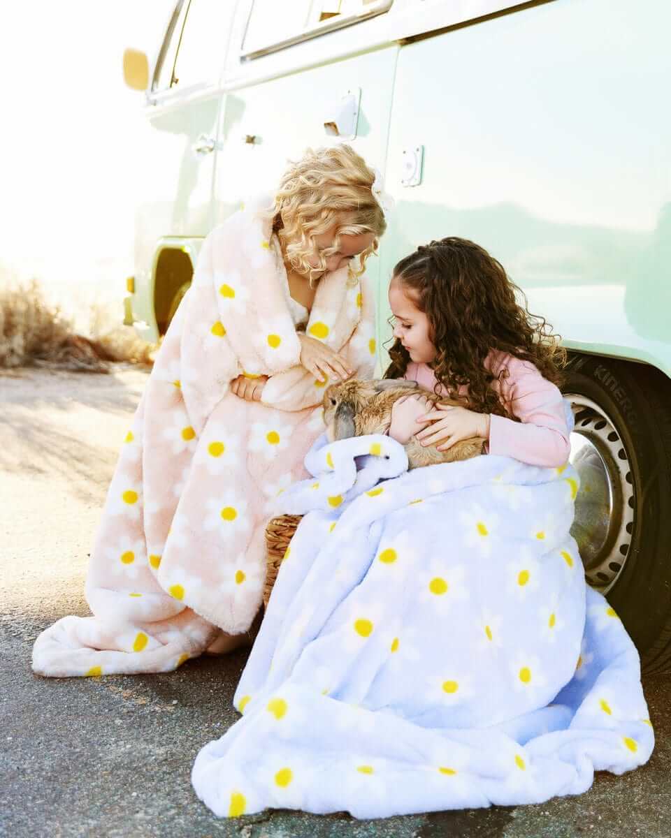 Two girls wrapped in Lush Flower Blankets in peach and lilac colors, with vibrant daisies against soft backgrounds.