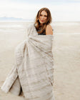 Woman wrapped in a Sorbet Tranquil Dove Blanket on a sandy beach, showcasing its soft texture and elegant color.