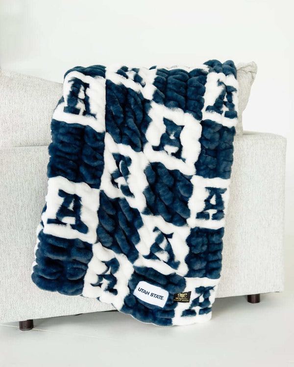 Navy and white checkered plush Utah State Aggies blanket draped over a light fabric sofa with logo tag visible.
