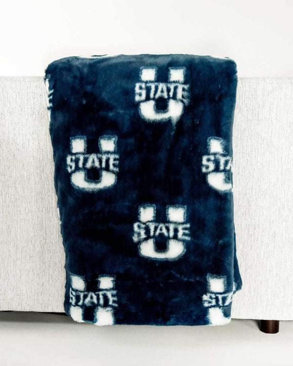 Navy Utah State Aggies plush throw blanket draped over light gray sofa with repeating State U logo pattern.