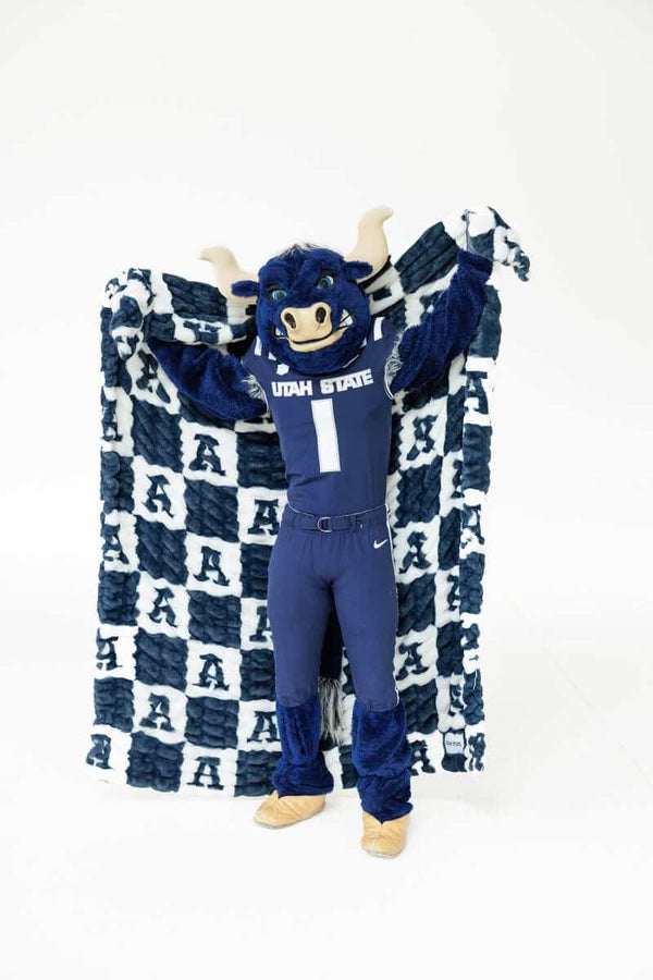 Utah State Aggies mascot holding a navy and white checkered blanket with large letter A pattern against a white studio background.