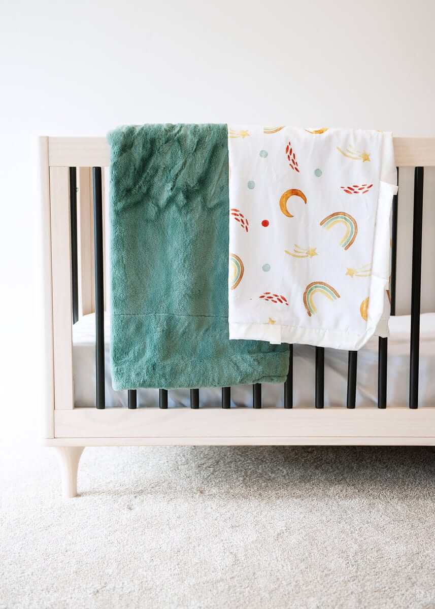 Thick mist green plush blanket and a rainbow, sun and moon themed blanket hanging over the edge of a crib.