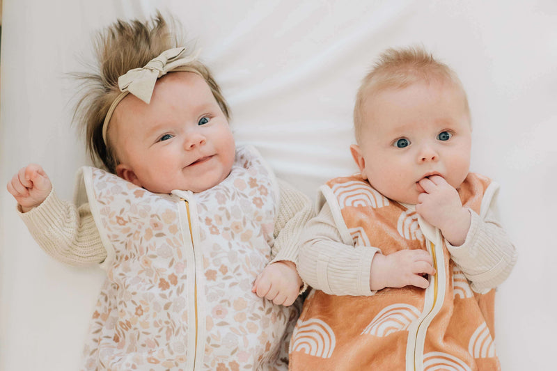 Two babies wearing Dainty Floral Wearable Minky Blanket and an orange rainbow design blanket, happily lying on a white surface.