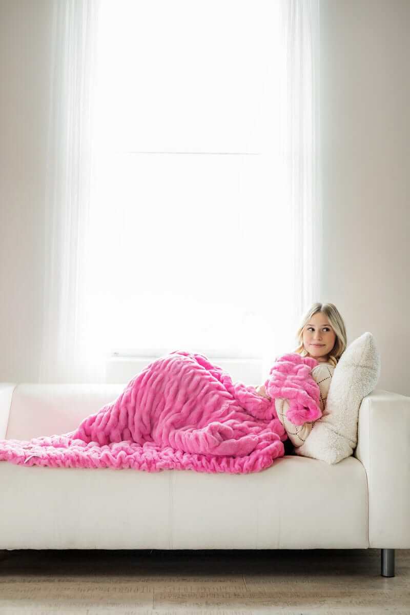 Young woman sitting on white couch snuggled up in a hot pink ruched plush blanket.
