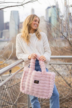 Woman showcasing the Annie Bag Ripple Blush while wearing a cozy sweater, set against a vibrant city backdrop.