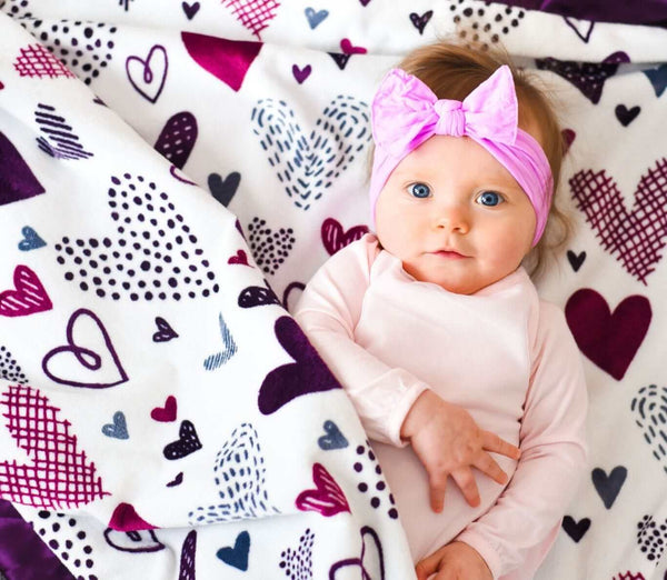 Baby girl cuddled in a Love Notes Plum Minky Couture blanket that has purple hearts of all sizes on a cream background.