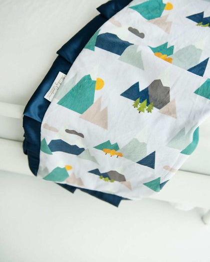 Blanket with mountaintops, trees and clouds in shades of misty green and neutral colors laid over the end of a bed. 
