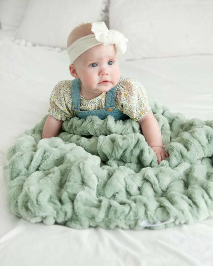 Baby girl sitting on a bed surrounded by a light green ruched mist blanket.