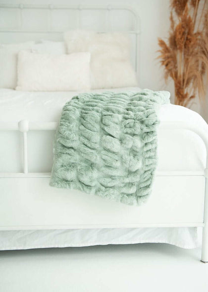 Hugs Mist Blanket draped over a white bed, showcasing its cozy sage green color and plush texture.