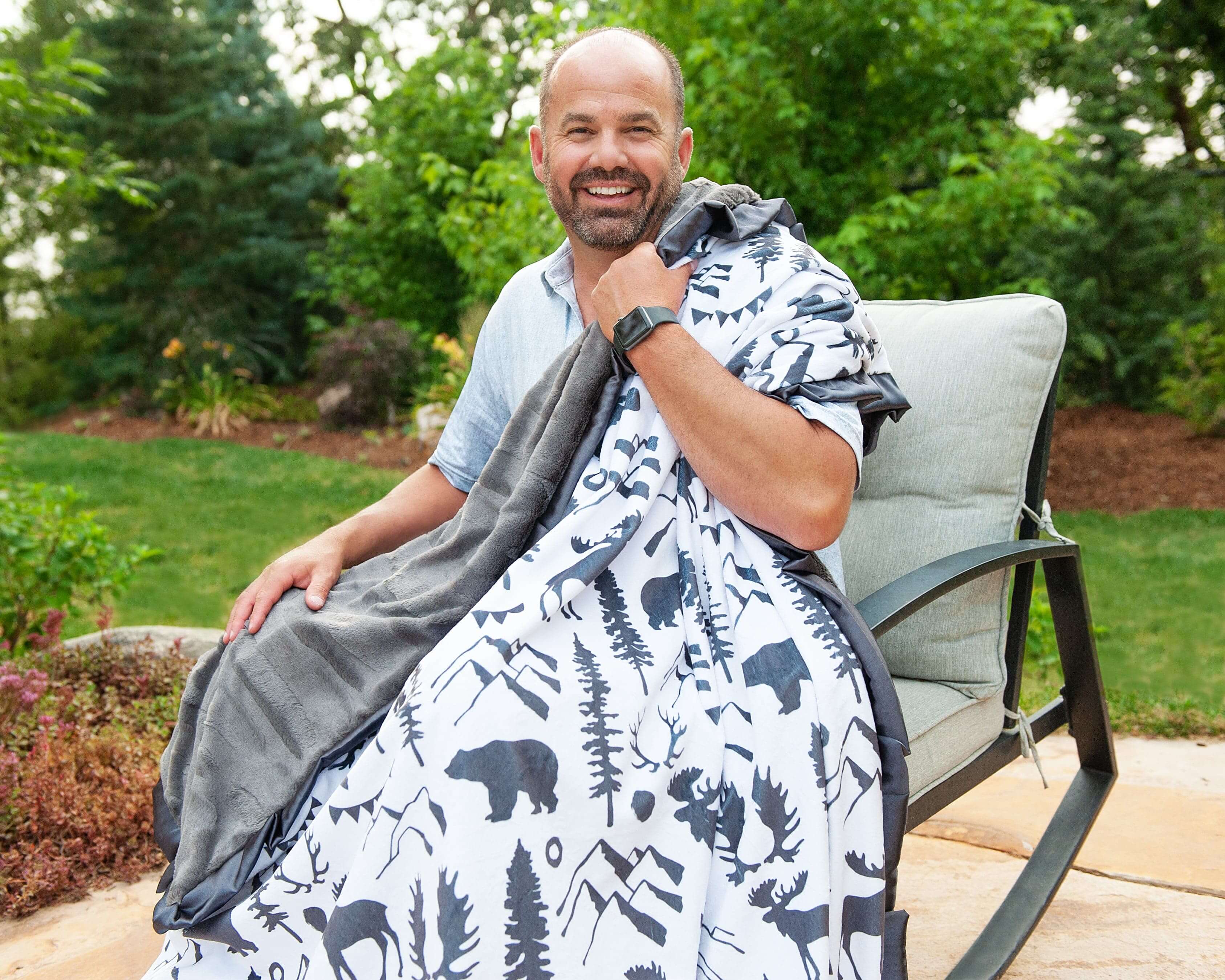 Smiling man wrapped in a Wilderness Blanket with wildlife designs, sitting outdoors in a cozy setting.
