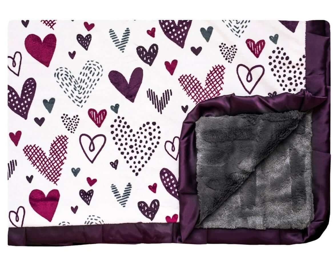 Love Notes Plum Minky Couture designer blanket laying flat with edge turned up. Doodled purple hearts with charcoal faux fur back and plum satin ruffle. 