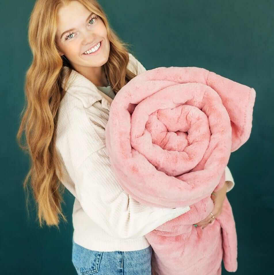 Woman smiling while holding a rolled up Lush Pretty Pink Blanket against a dark green background.