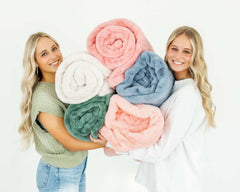 Two women holding a stack of colorful Lush Pretty Pink Blankets, showcasing soft textures and pastel hues.
