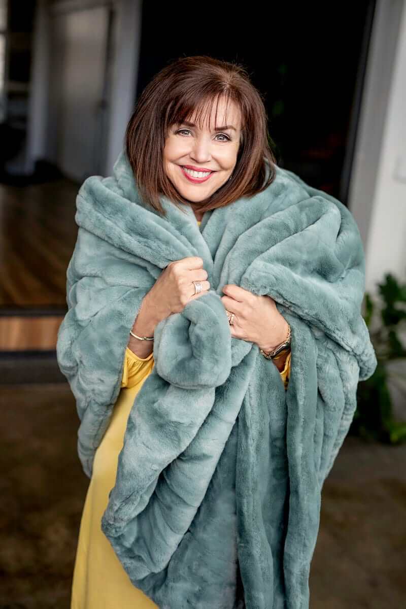 A woman wrapped in a Lush Midnight Mist Blanket, showcasing its thick misty green plush fabric and cozy texture.
